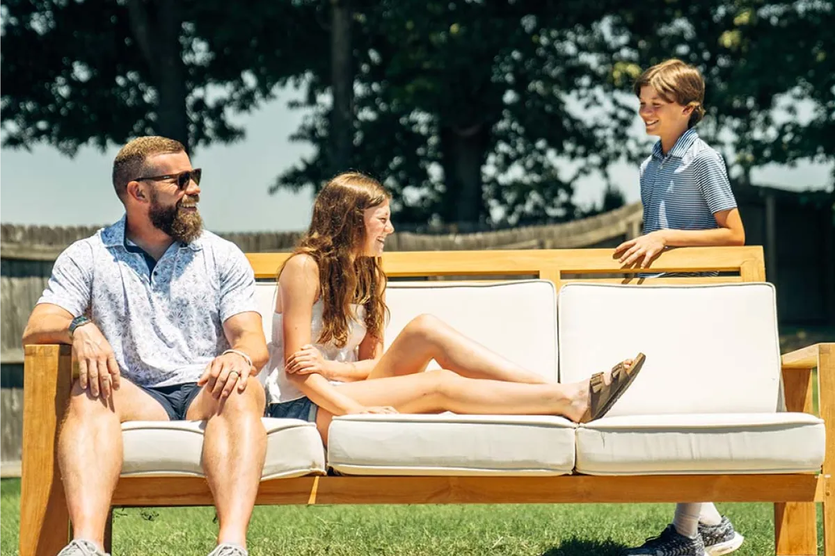 Family relaxing on a teak outdoor sofa with white cushions in a sunny backyard.